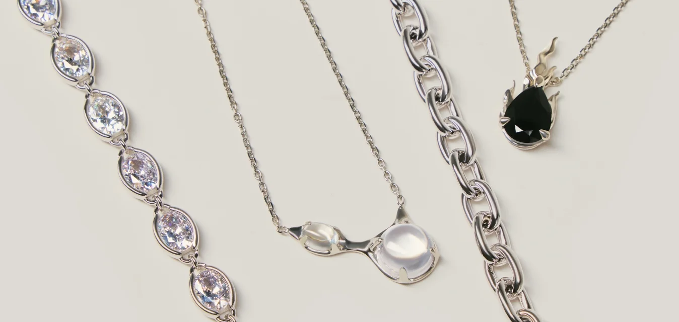 Cinque collane e catene d'argento su uno sfondo beige, con pietre preziose a forma di goccia trasparenti e nere e design a catena collegata.
