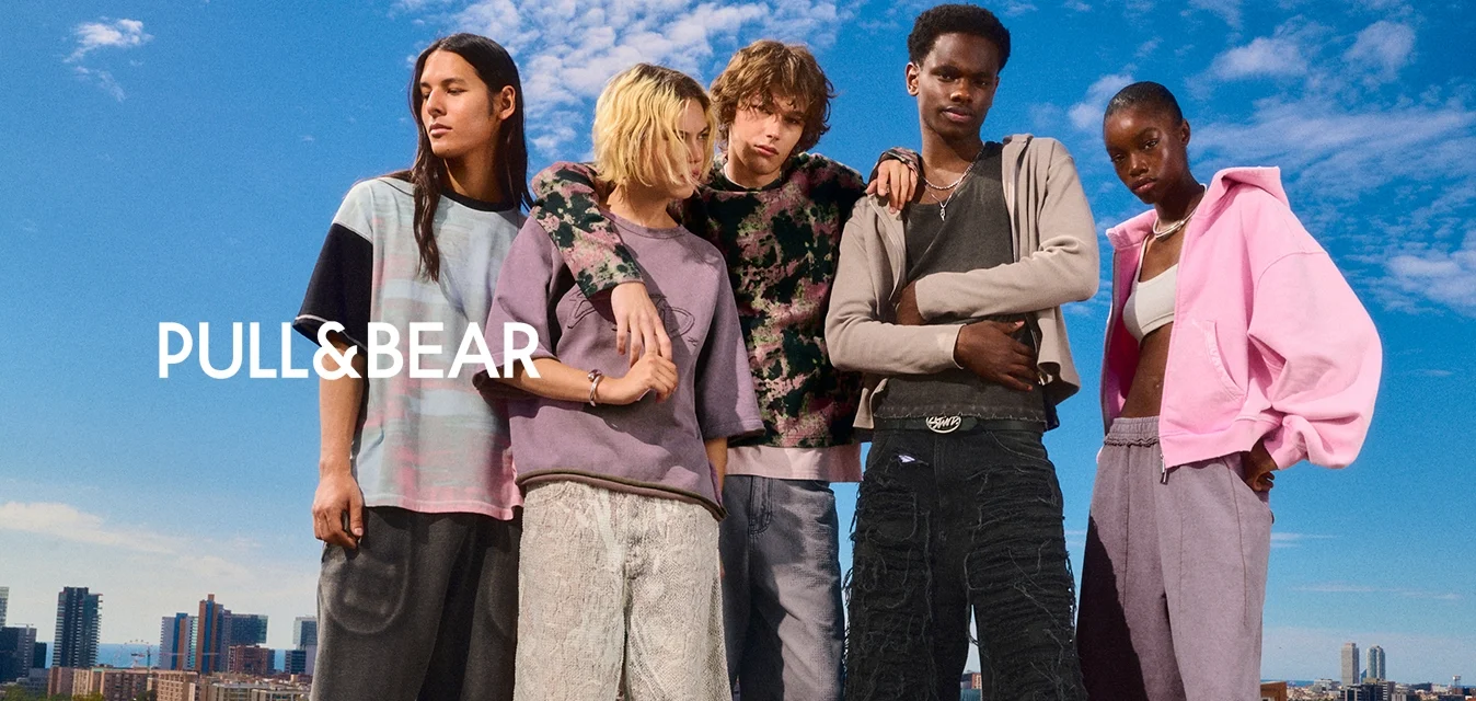 Cinque giovani adulti che indossano abiti larghi e casual e pantaloni corti si trovano vicini l'uno all'altro davanti a uno skyline cittadino sotto un cielo blu.