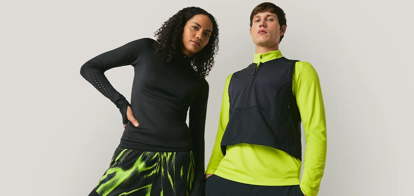 Due modelli posano contro uno sfondo semplice, uno indossa abbigliamento sportivo nero e verde neon, l'altro una maglia nera con giacca verde neon e giubbotto.