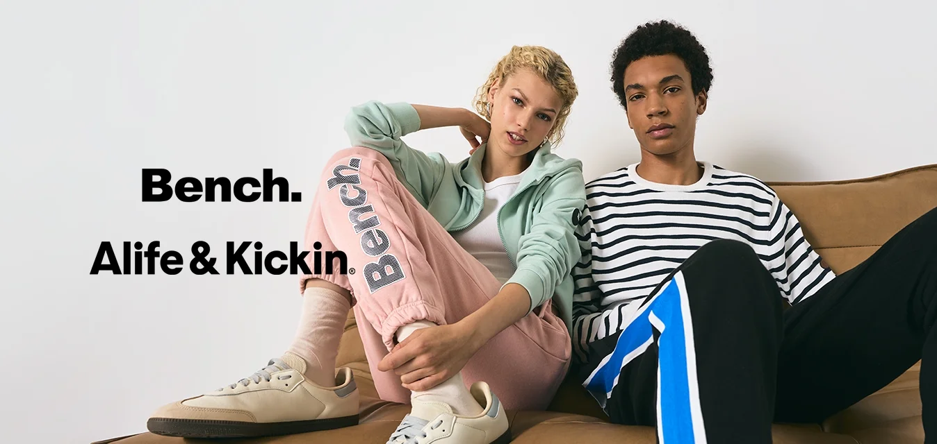 Due giovani adulti seduti su un divano in pelle marrone, uno che indossa pantaloni sportivi rosa pastello e una giacca verde chiaro, l'altro con una maglietta a righe e pantaloni neri.