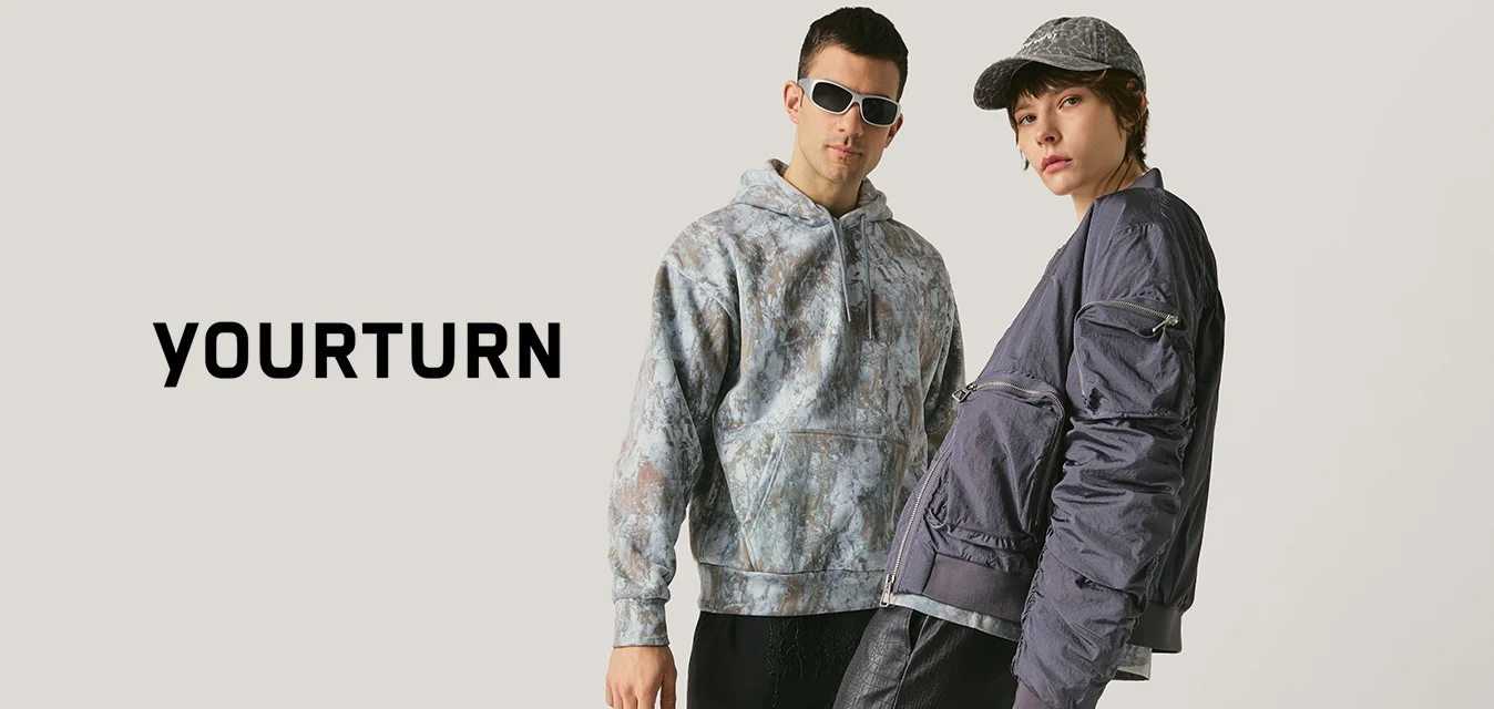 Due persone vestite alla moda posano davanti a uno sfondo neutro, indossando pantaloni scuri, top a strati e accessori come occhiali da sole e una borsa.