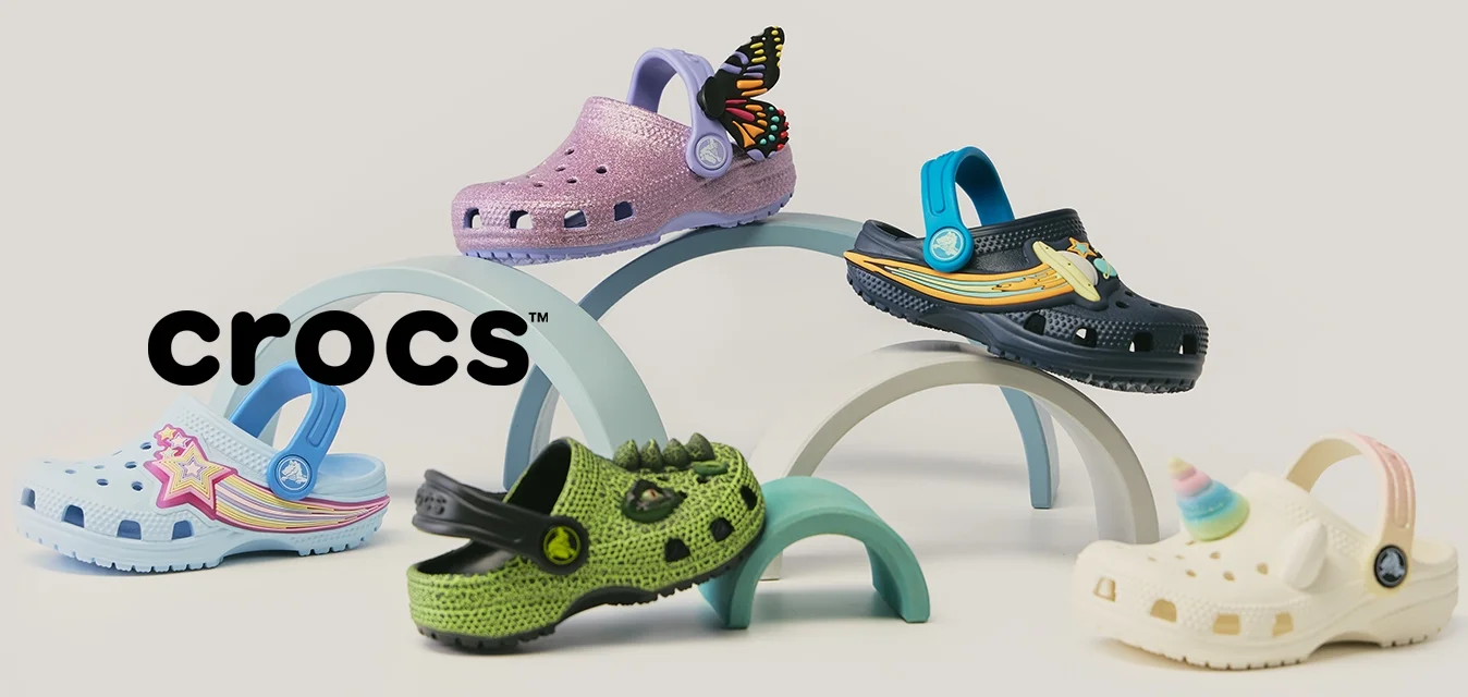 Cinque zoccoli colorati per bambini con charm decorativi disposti su archi pastello contro uno sfondo neutro.