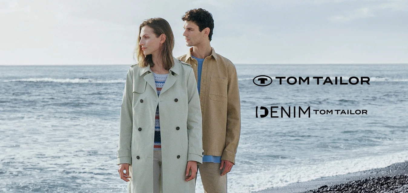 Trench coat verde chiaro, camicia beige, pantaloni bianchi e scarpe marroni. Entrambi gli abiti sono casual, con tessuti morbidi contro uno sfondo di spiaggia rocciosa.