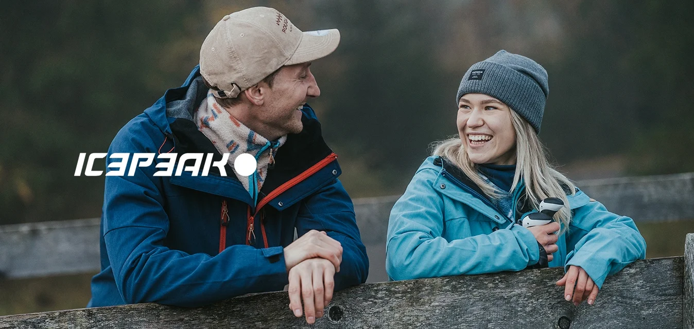 Due persone si appoggiano a una recinzione di legno. Una indossa una giacca blu navy con dettagli rossi, e l'altra una giacca azzurro chiaro e un berretto grigio.
