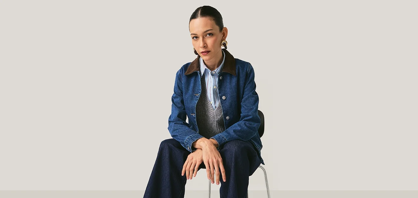 Giacca di jeans con colletto marrone, gilet grigio e camicia a righe azzurro chiaro. Jeans scuri a gamba larga e scarpe marroni con lacci. Seduto su una sedia.