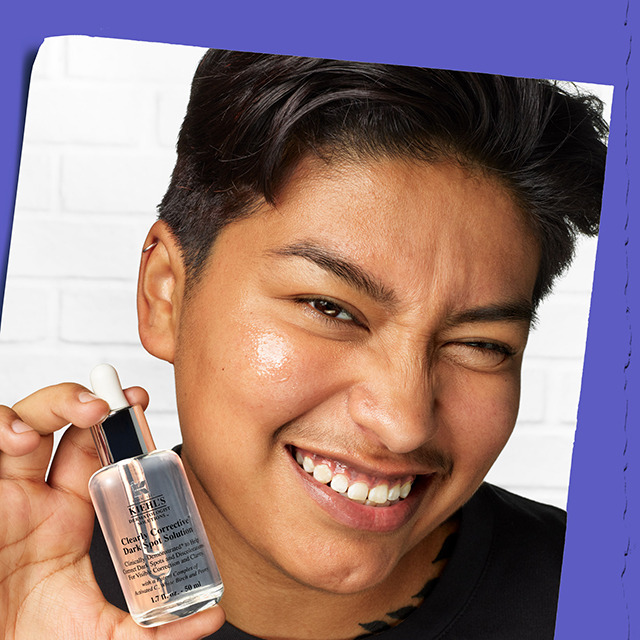 Un ragazzo sorridente mostra la boccetta di siero Clearly Corrective Dark Spot Solution di Kiehl's vicino al viso, evidenziando una pelle luminosa su uno sfondo viola vibrante.