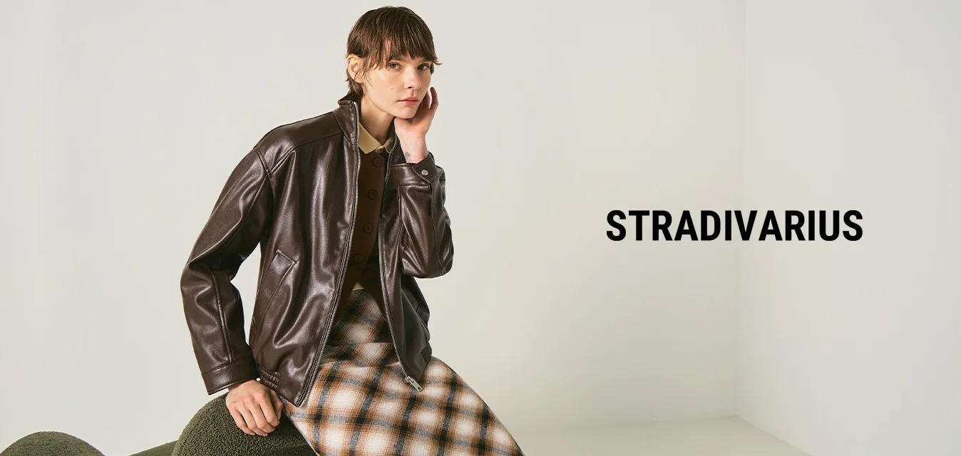 Persona con giacca di pelle marrone scuro e gonna a quadri, seduta su una sedia verde dalla texture evidente contro uno sfondo chiaro uniforme.