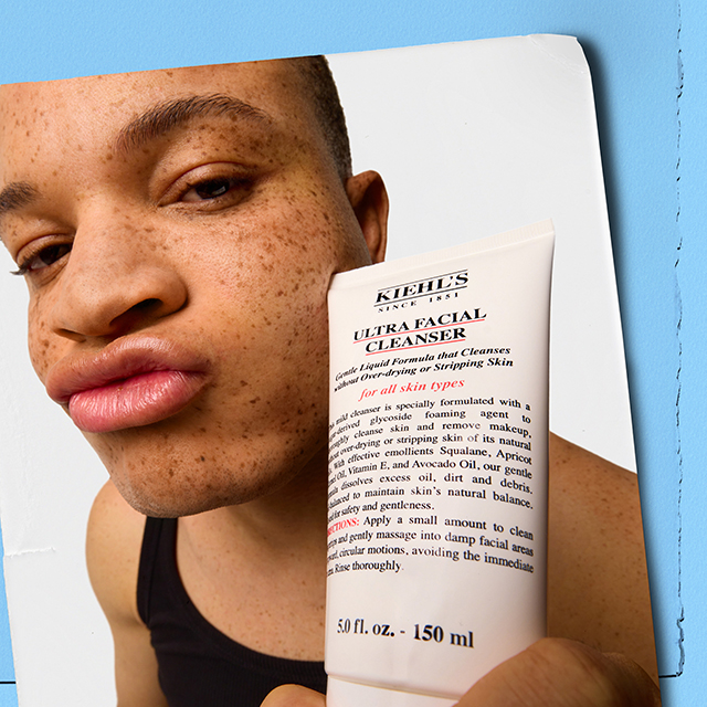 Primo piano di un ragazzo con lentiggini che tiene in mano il tubo di Ultra Facial Cleanser di Kiehl's, su uno sfondo azzurro chiaro in stile collage fotografico.