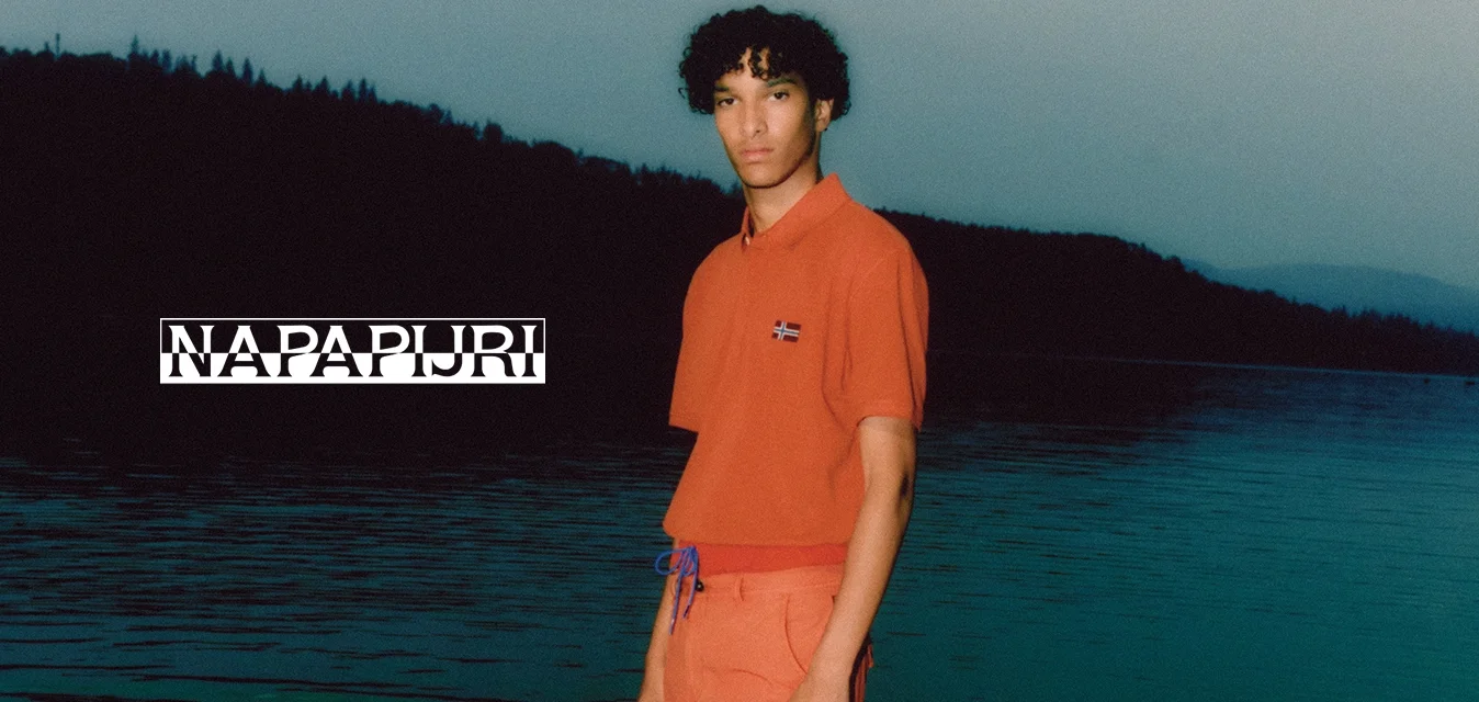 Polo arancione a maniche corte con un piccolo dettaglio della bandiera, abbinato a pantaloni corti arancioni coordinati con coulisse. Acqua sullo sfondo.