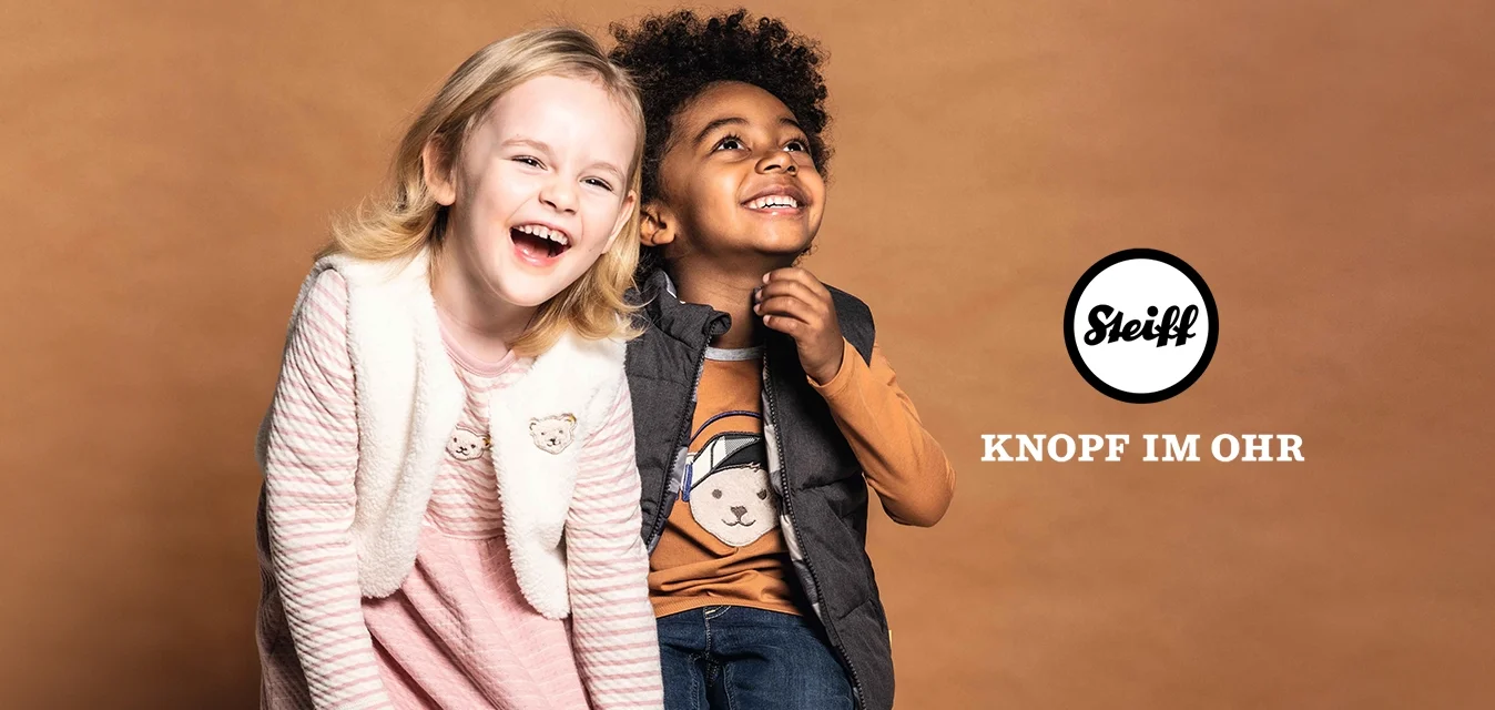 Due bambini sono seduti su una cassa. La ragazza indossa un vestito a righe rosa e un gilet crema; il ragazzo indossa un gilet grigio sopra una maglietta con una grafica di un orso marrone e jeans.