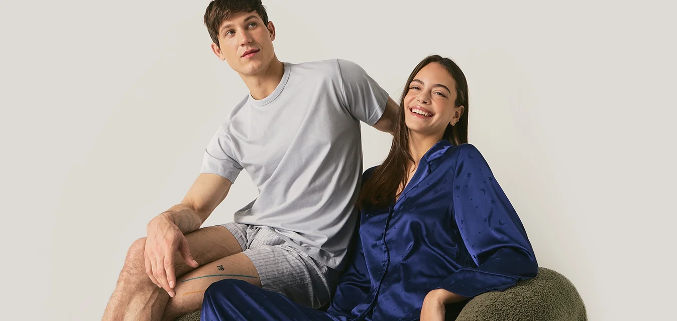 Giovane uomo in pigiama grigio e giovane donna in pigiama di seta blu seduti su un divano verde su sfondo semplice.