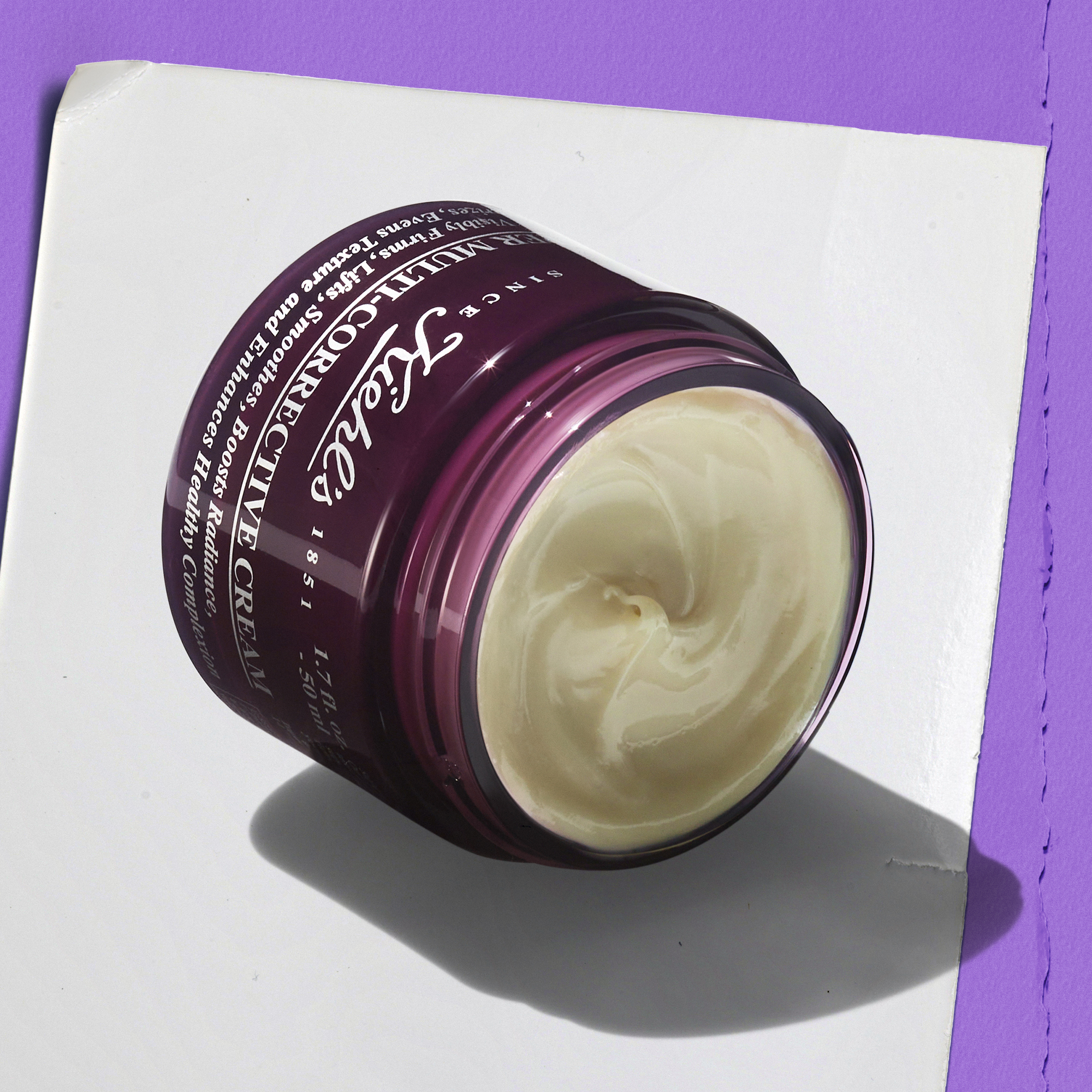 Vasetto aperto di Super Multi-Corrective Cream Kiehl&rsquo;s in confezione viola inclinato su superficie chiara; la crema beige dalla texture ricca &egrave; visibile all&rsquo;interno. Luce viola laterale con ombra definita.