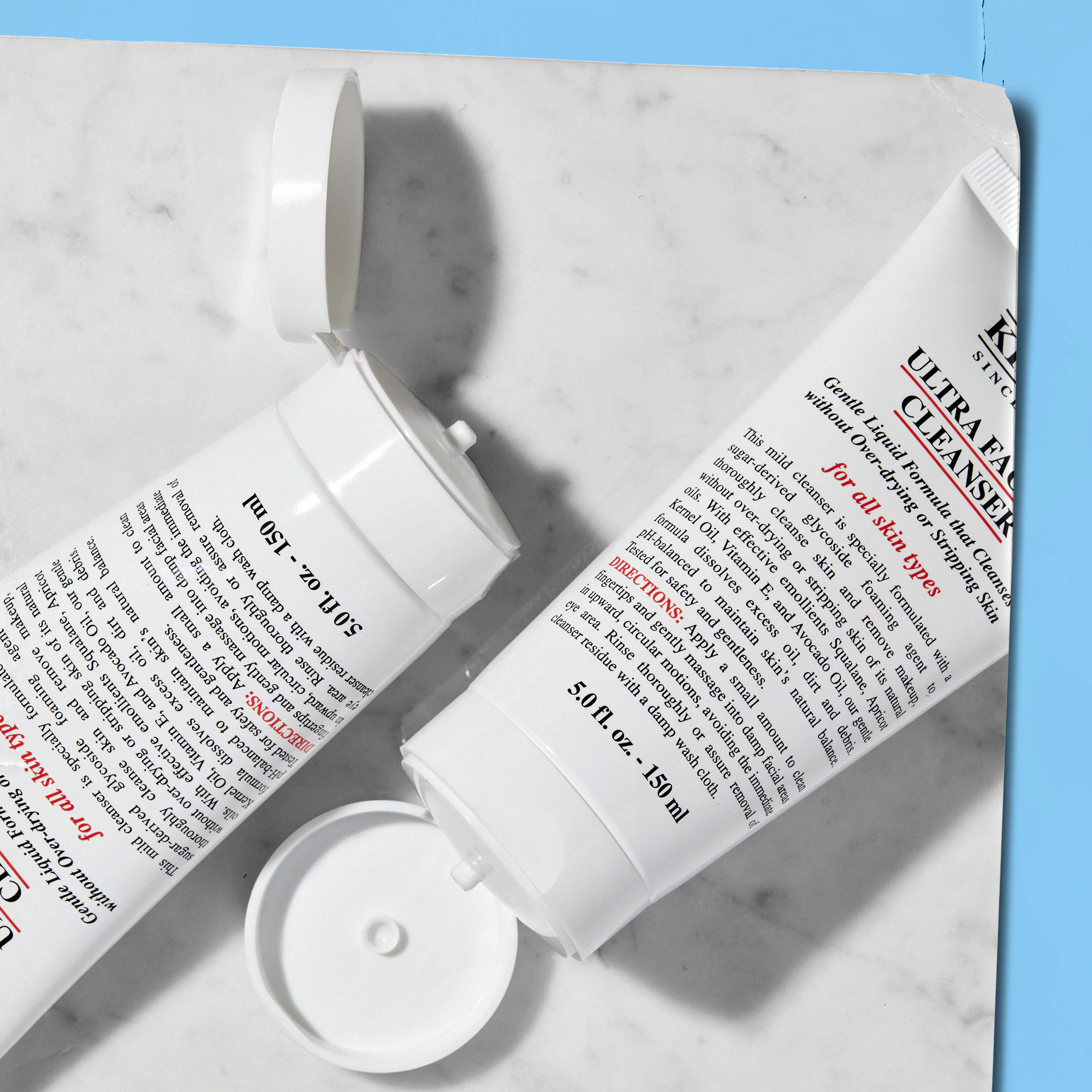 Due tubi di Ultra Facial Cleanser Kiehl&rsquo;s da 150 ml appoggiati su superficie effetto marmo; uno &egrave; aperto con tappo ribaltato. Sfondo con luce blu laterale e ombre morbide.