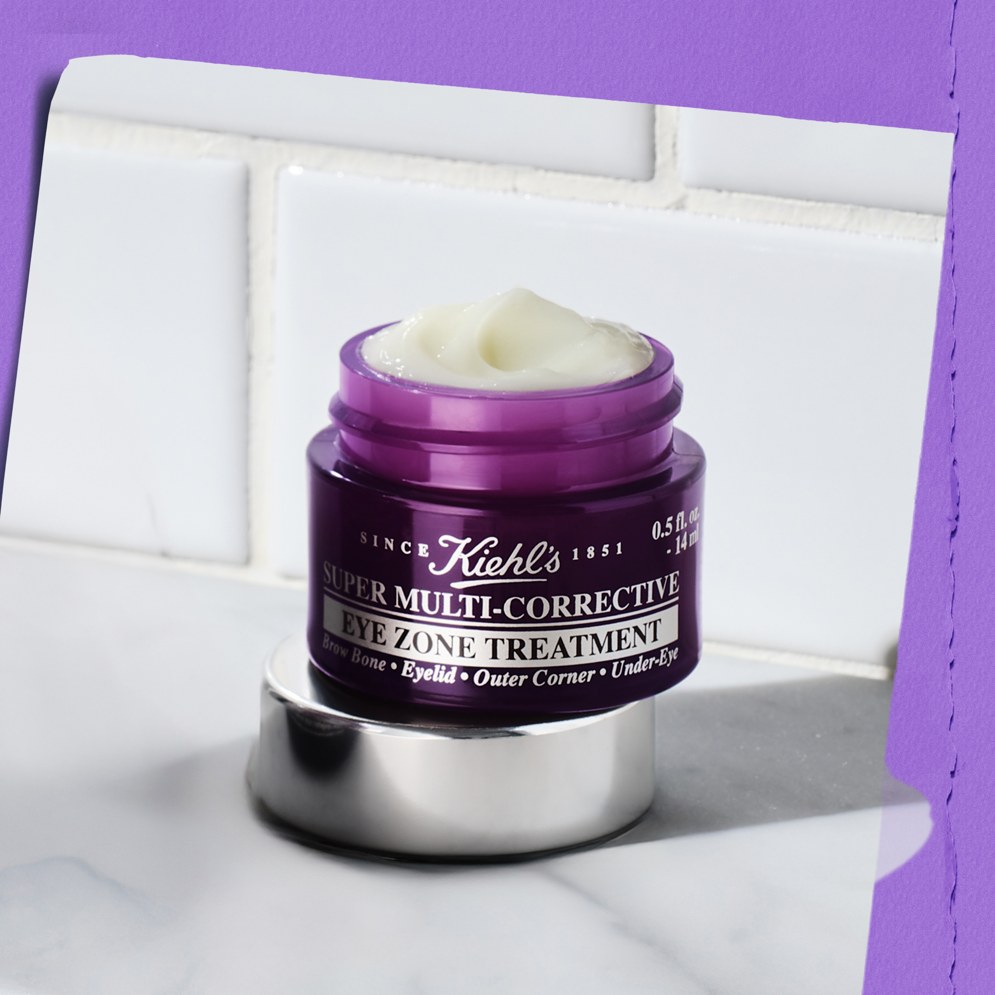 Vasetto aperto di Super Multi-Corrective Eye Zone Treatment Kiehl&rsquo;s in confezione viola, con crema color avorio visibile. Il prodotto &egrave; posizionato su base argentata davanti a piastrelle bianche, luce viola laterale.