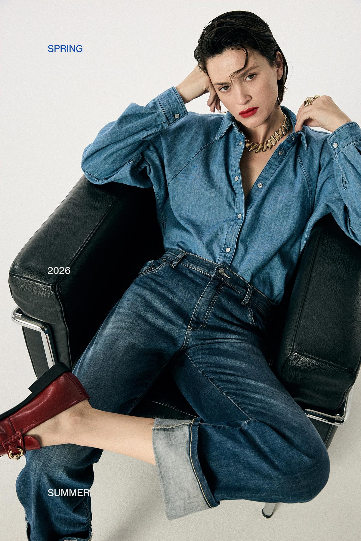 Model reclining in black armchair wearing denim shirt and rolled jeans with red loafers and gold chain, Spring/Summer 2026 fashion