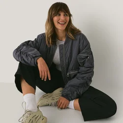 Giovane donna in abbigliamento casual e sneakers seduta sul pavimento contro un muro bianco semplice, sorridente e che guarda di lato.