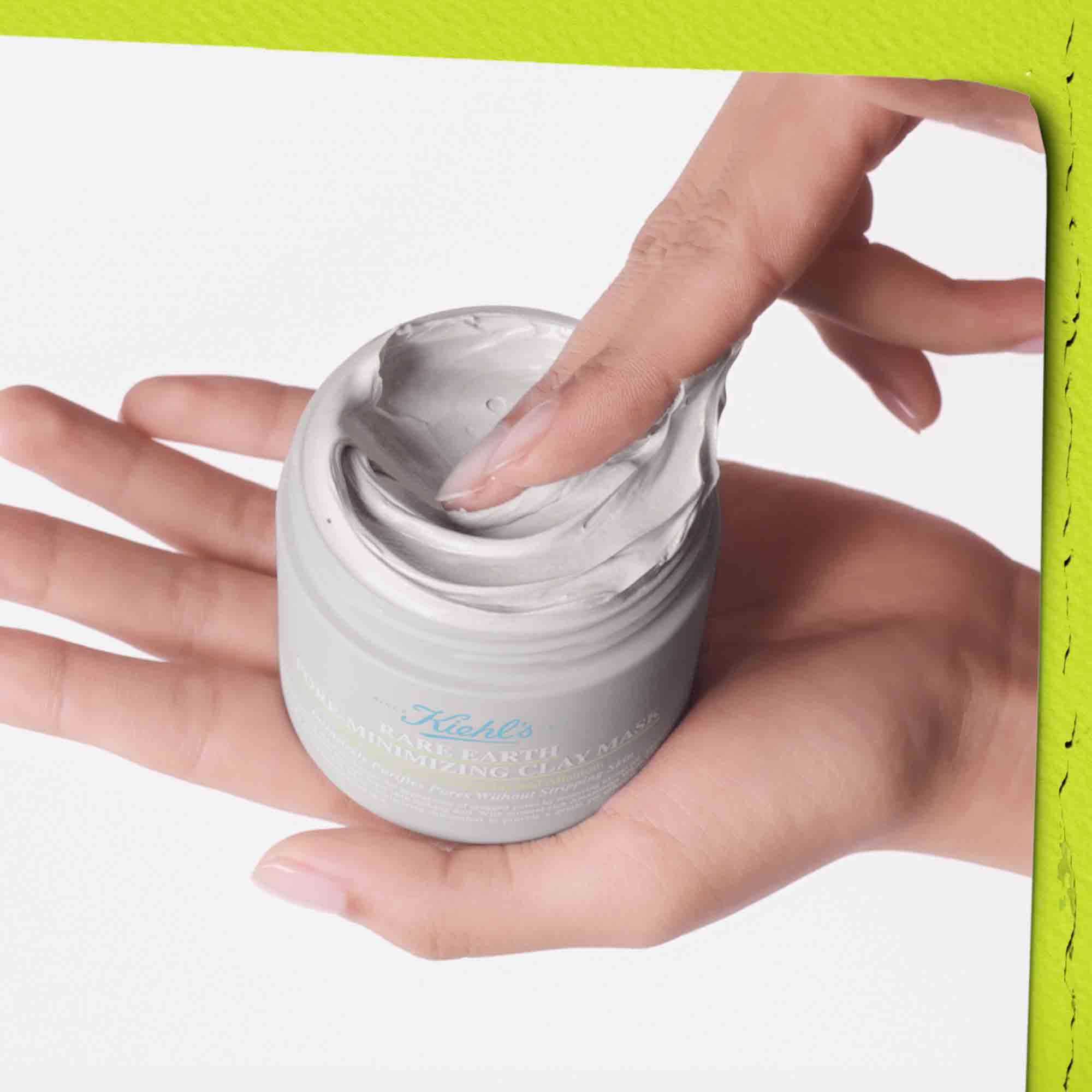 Barattolo aperto di Rare Earth Deep Pore Cleansing Masque Kiehl&rsquo;s appoggiato su una mano; una seconda mano preleva la maschera grigia dalla texture densa e cremosa. Sfondo neutro con bordo verde lime.