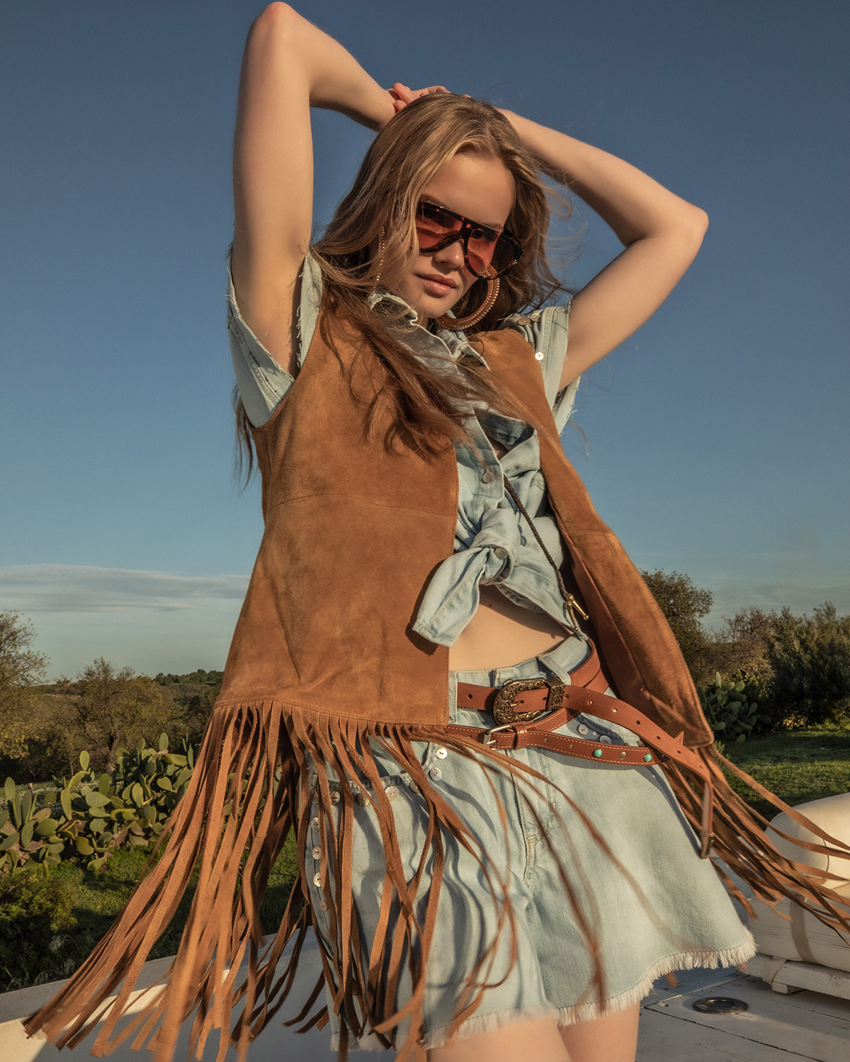 Woman wearing suede fringe vest, knotted denim shirt, mini denim skirt and sunglasses posing outdoors in western-inspired fashion