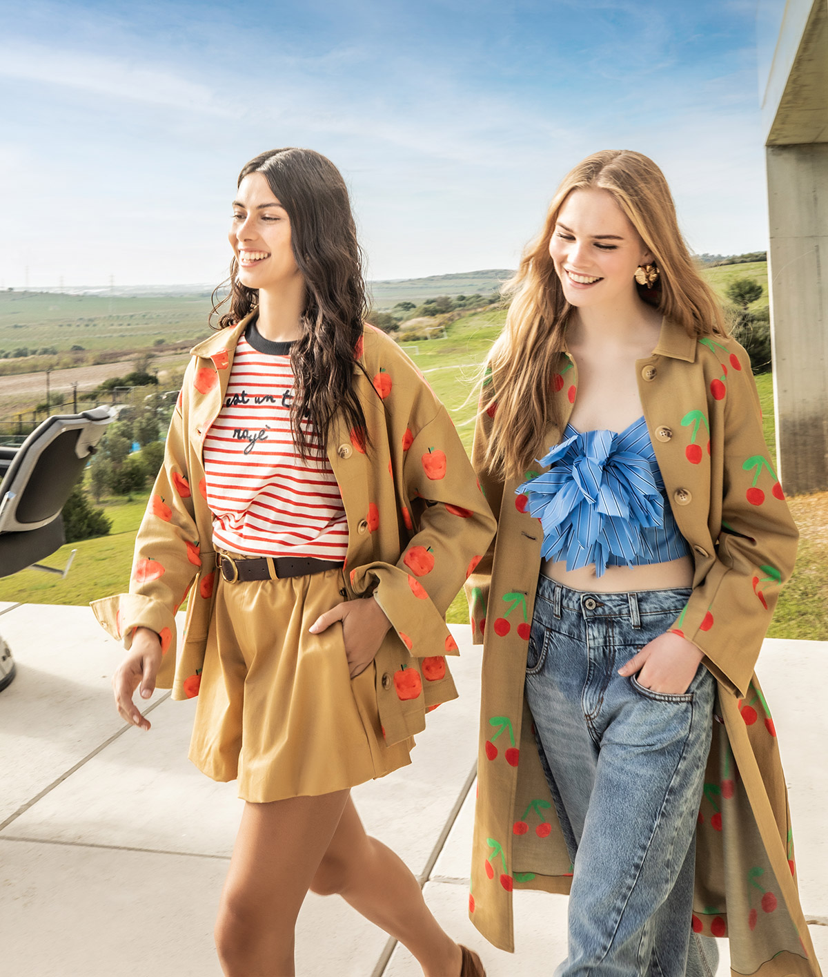Two smiling women walking outdoors in cherry-print coats and stylish spring outfits against a sunny countryside backdrop
