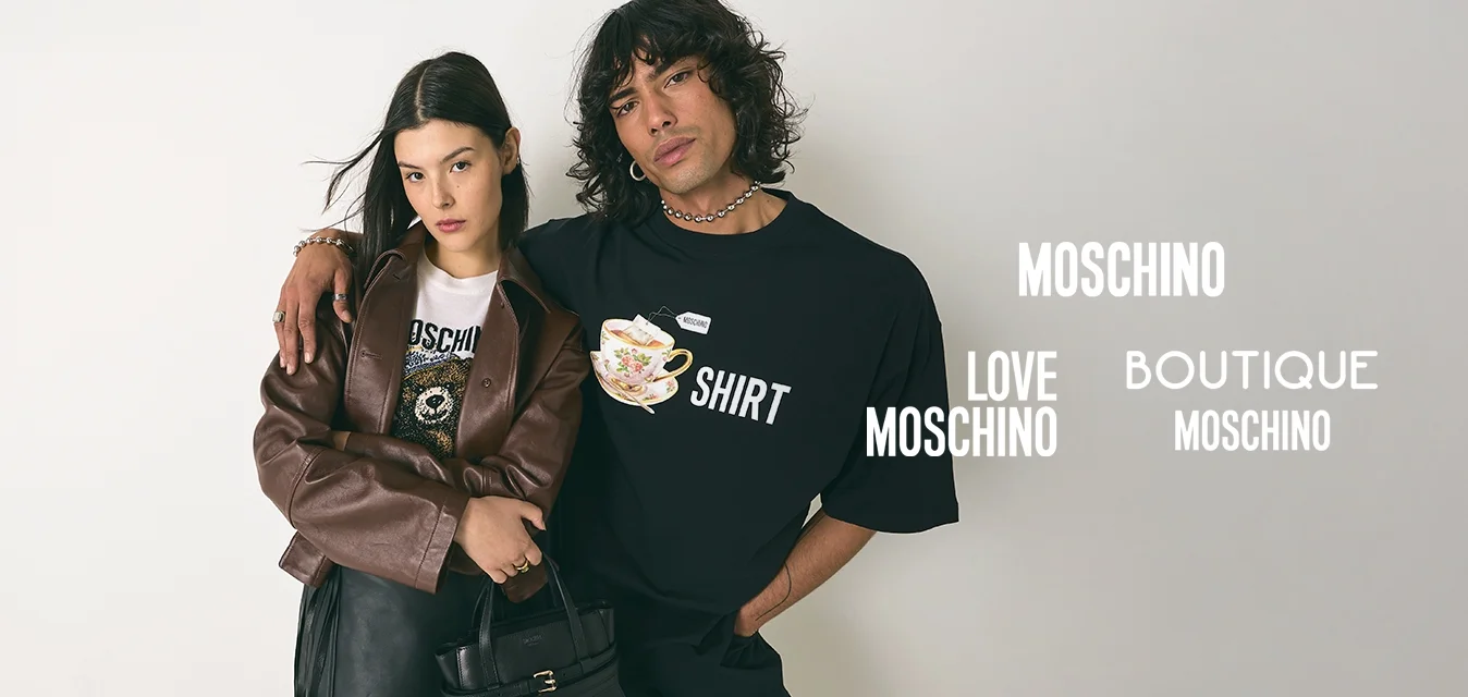 Giovane con capelli ricci in una maglietta nera con grafica ha un braccio attorno a una donna che indossa una giacca marrone, una gonna nera e tiene una borsa nera.