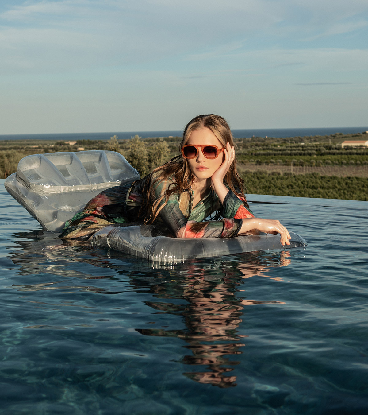 Young woman reclining on an inflatable float in an infinity pool, wearing sunglasses and a colourful dress with sea and countryside horizon