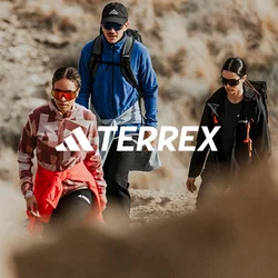 Tre escursionisti con abbigliamento da esterno e occhiali da sole camminano su un sentiero roccioso in un paesaggio secco e erboso.