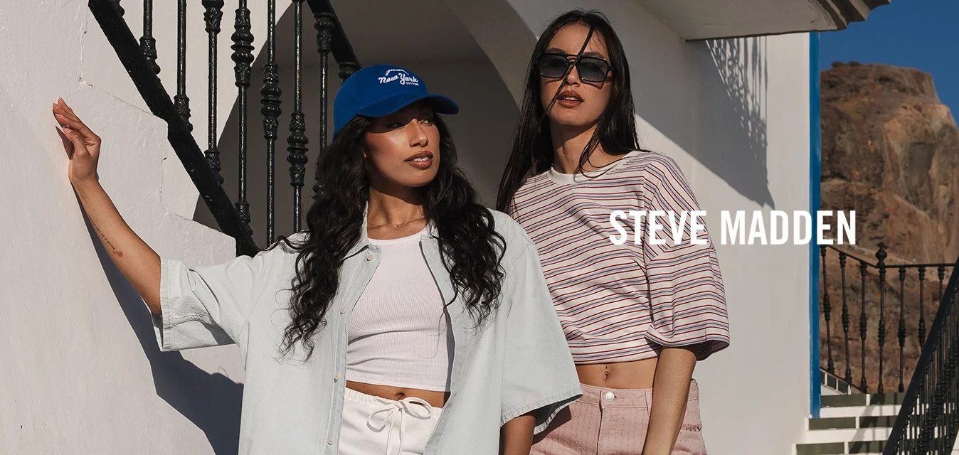 Due donne con abiti estivi casual posano vicino a un edificio bianco con rifiniture blu, una scala esterna e una costa rocciosa sotto un cielo sereno.