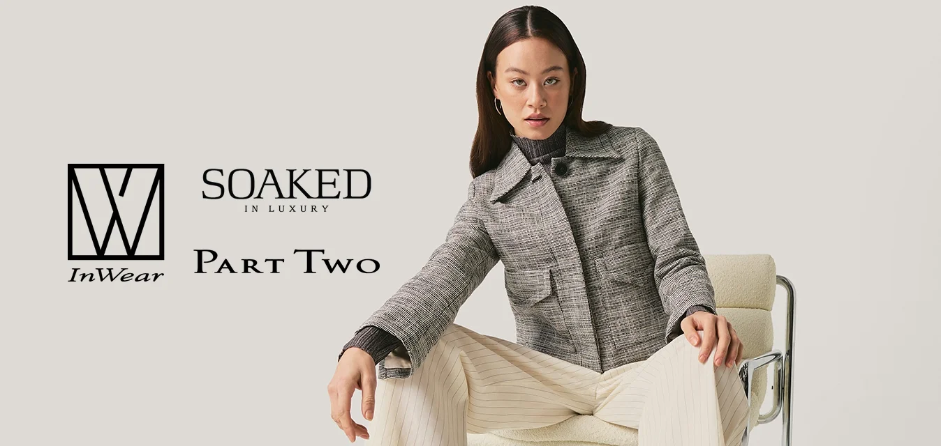 Donna con capelli neri che indossa una giacca grigia e pantaloni crema è seduta su una sedia con struttura in metallo su sfondo bianco uniforme.