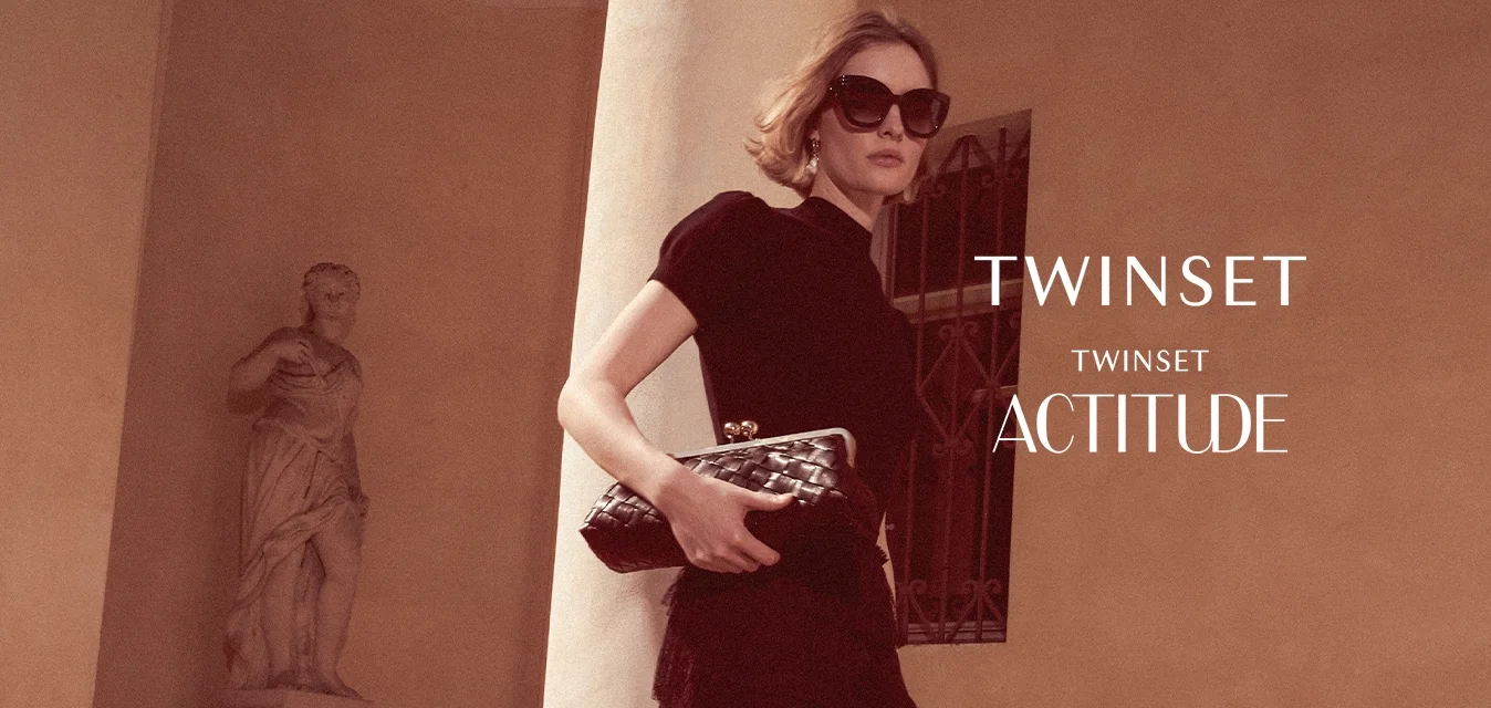 Donna con vestito nero e occhiali da sole tiene una pochette mentre cammina su gradini di pietra accanto a una colonna e una statua classica.