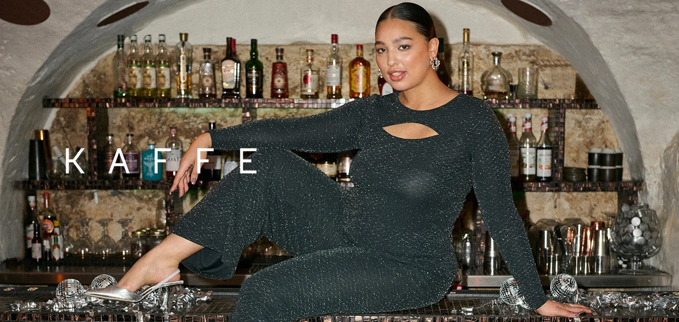 Catsuit nero lucido con maniche lunghe e dettaglio a taglio, abbinato a ballerine argento. Sullo sfondo si vede un bar con bottiglie.