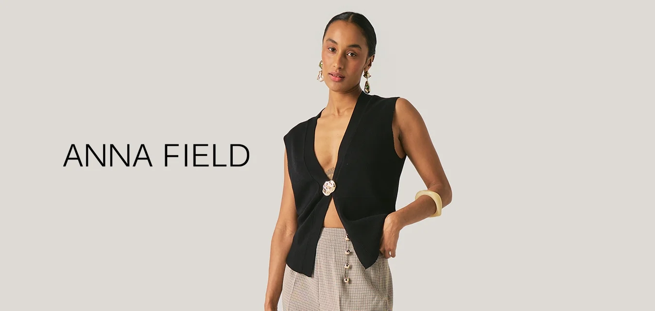 Donna che indossa un top nero senza maniche con chiusura frontale, pantaloni beige ampi con fantasia, scarpe nere, orecchini a cerchio e tiene in mano una piccola borsa dorata.