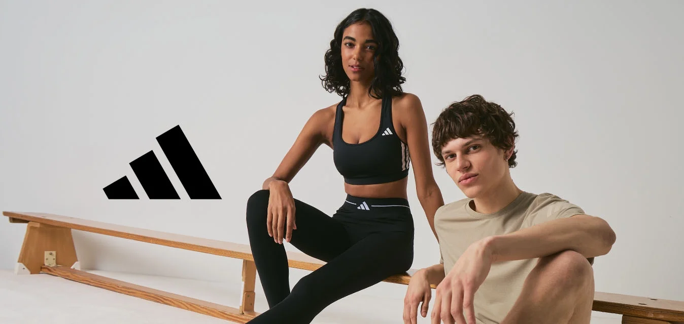 Due giovani adulti in abbigliamento sportivo sono seduti vicino a panche di legno in uno spazio interno minimalista, entrambi indossano scarpe da ginnastica Adidas.