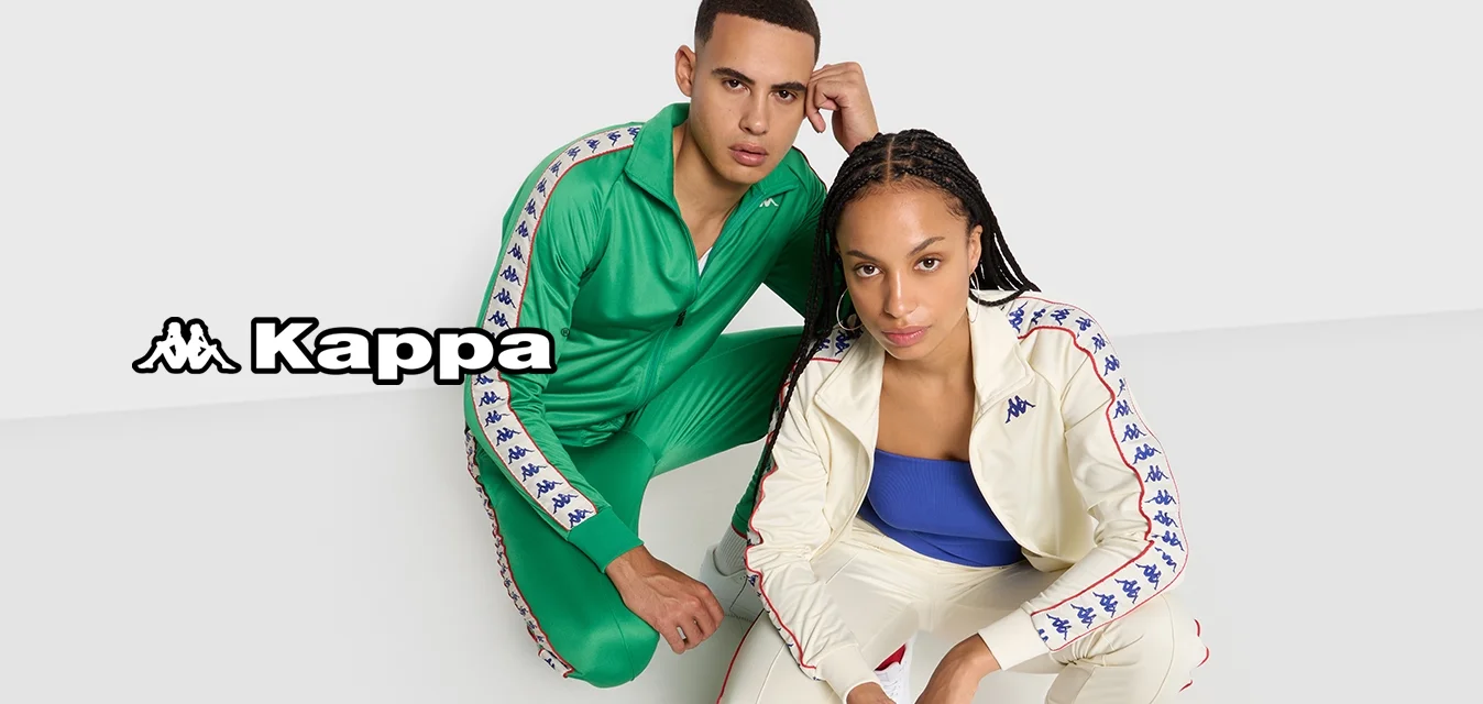 Due persone si accuattano su un pavimento bianco puro, indossando tute sportive coordinate: una verde e l'altra crema con un top blu e sneaker bianche.