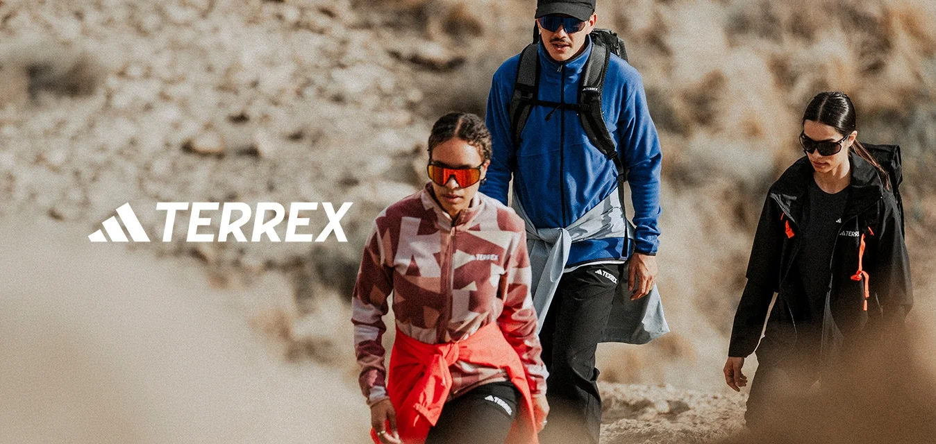 Tre escursionisti con abbigliamento da esterno e occhiali da sole camminano su un sentiero roccioso in un paesaggio secco e erboso.