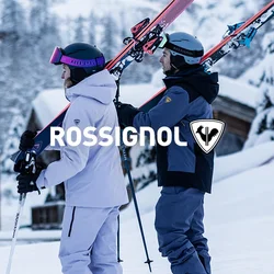 Quattro sciatori in abbigliamento invernale portano gli sci sulle spalle, in piedi sulla neve vicino a baite di legno con montagne innevate sullo sfondo.