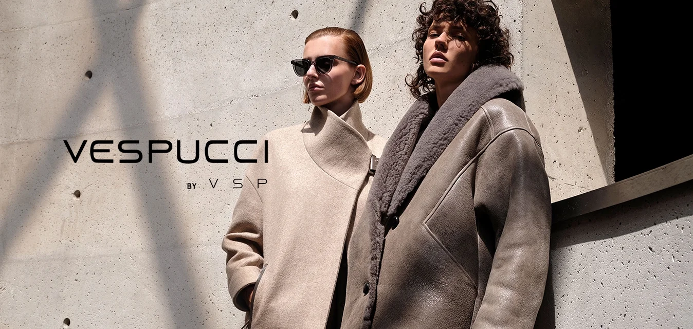 Due donne con eleganti cappotti invernali oversize si trovano contro un muro di cemento testurizzato con ombre che proiettano linee.