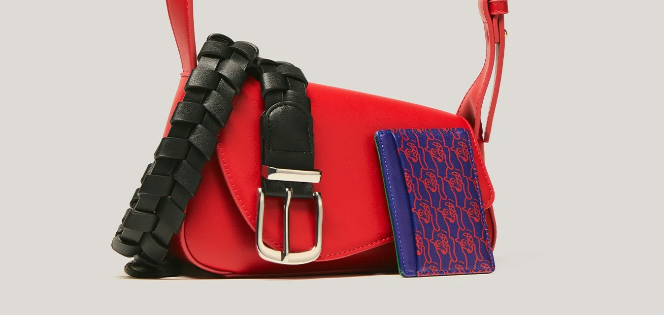 Borsa a tracolla rossa con cinturino regolabile, cintura nera intrecciata con fibbia d'argento e piccolo portafoglio blu con motivo su sfondo neutro.
