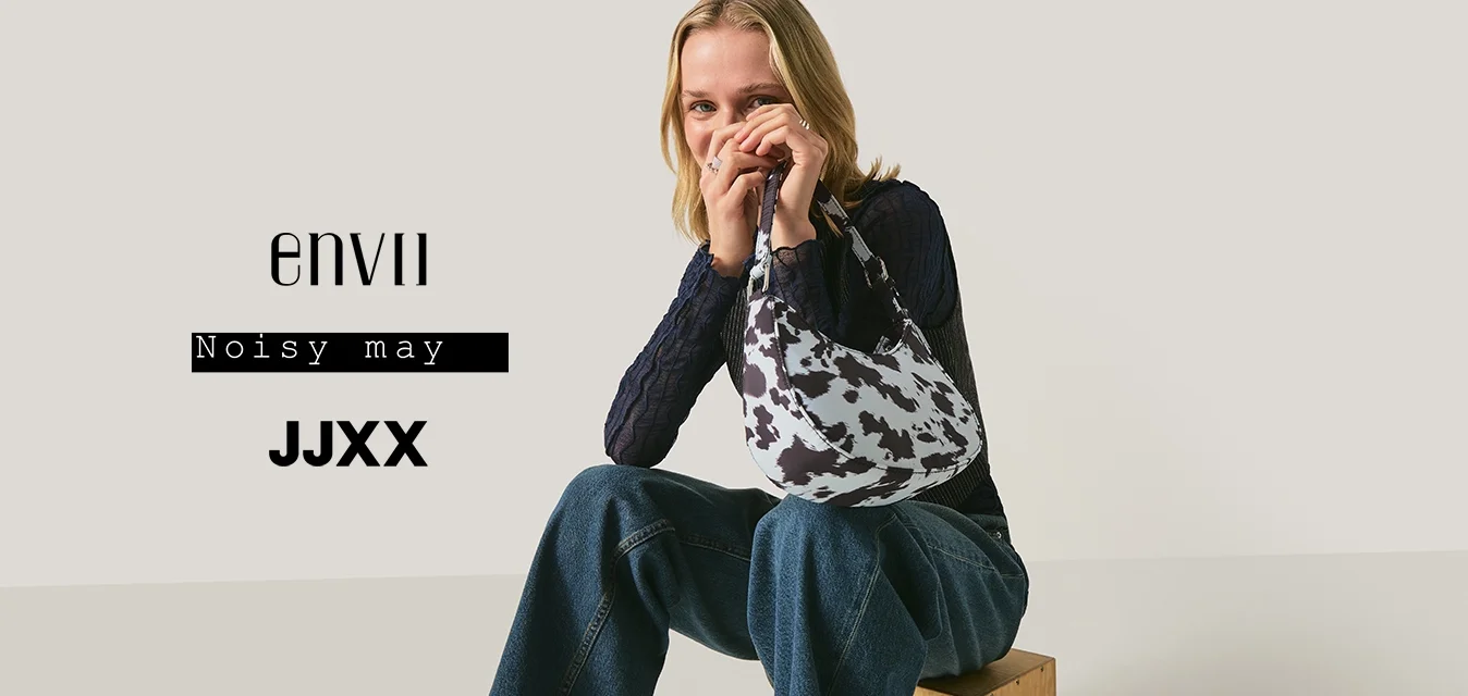 Giovane donna seduta su una cassa di legno che tiene in mano una borsa con stampa a mucca bianca e nera, indossa un top in pizzo blu navy e jeans a gamba larga.