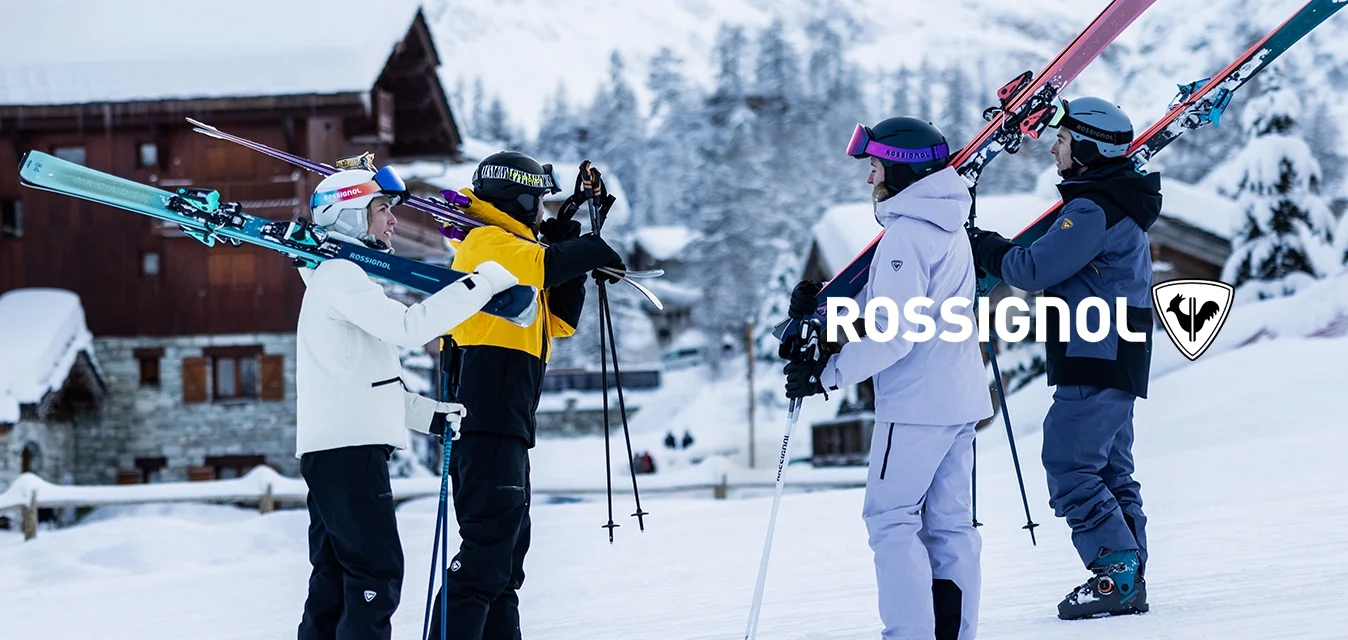 Quattro sciatori in abbigliamento invernale portano gli sci sulle spalle, in piedi sulla neve vicino a baite di legno con montagne innevate sullo sfondo.