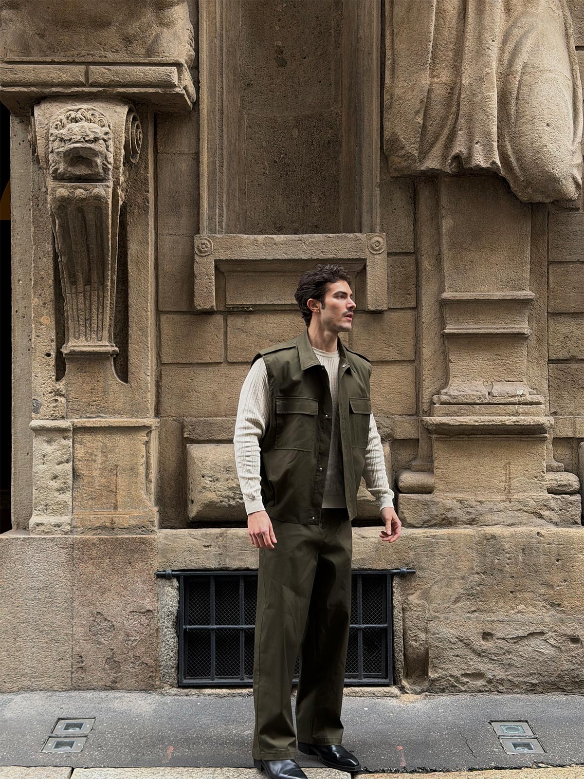 Man in olive utility vest and trousers over cream knit sweater standing by ornate stone building facade