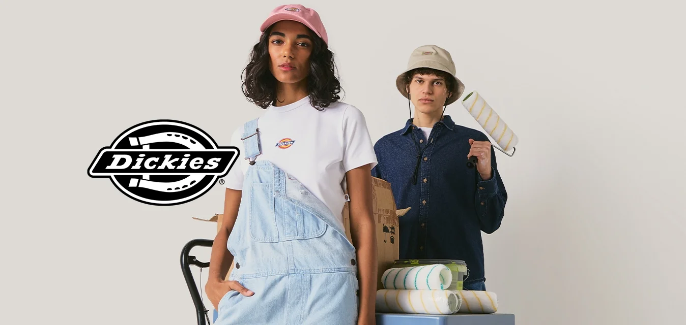 Giovane donna con cappellino rosa e tuta che tiene un pennello, giovane uomo con cappello a secchiello con rullo per pittura in piedi accanto a un tavolo con materiali.