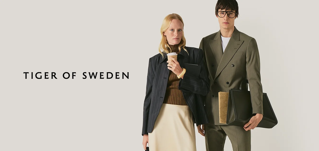 Due persone vestite professionalmente in piedi: donna con gonna beige, stivali neri, che tiene un caffè e una borsa; uomo con completo oliva che tiene una grande borsa nera.