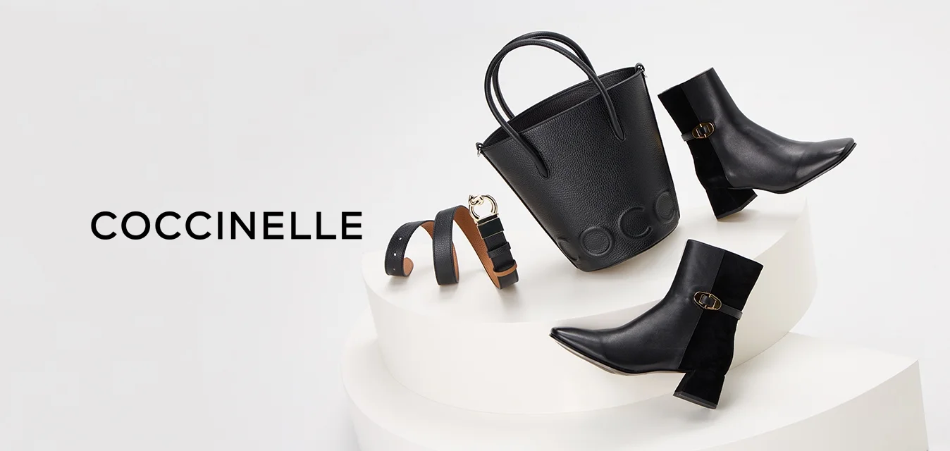 Borsa a secchiello in pelle nera con doppi manici, accompagnata da stivali neri alla caviglia con punta quadrata e dettaglio in fibbia dorata, su un piedistallo bianco.