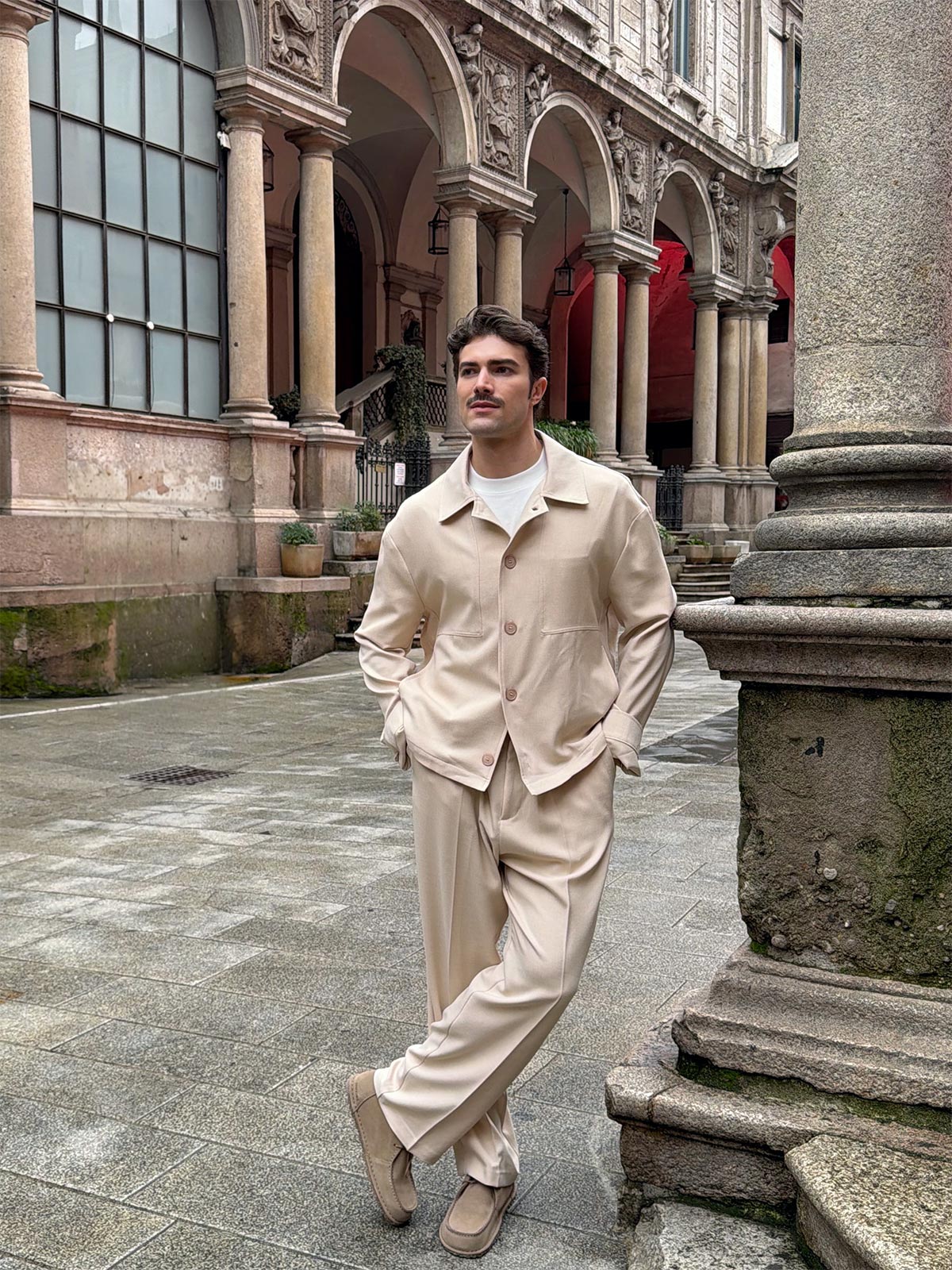 Man in beige relaxed suit and trainers leaning against stone column in historic arched courtyard, stylish men's outfit