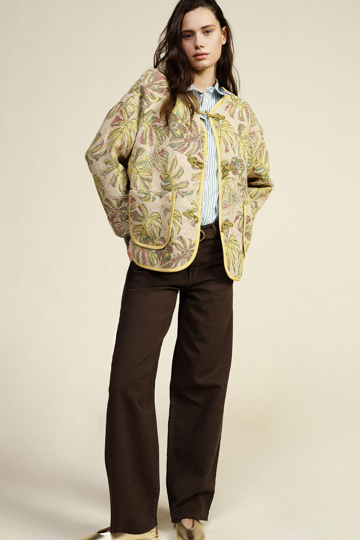 Woman wearing embroidered quilted floral jacket over a striped shirt with brown wide‑leg trousers, hands in pockets on neutral background