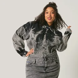 Donna con capelli ricci che indossa una giacca e una gonna di jeans nero e grigio, in piedi davanti a uno sfondo chiaro e uniforme.