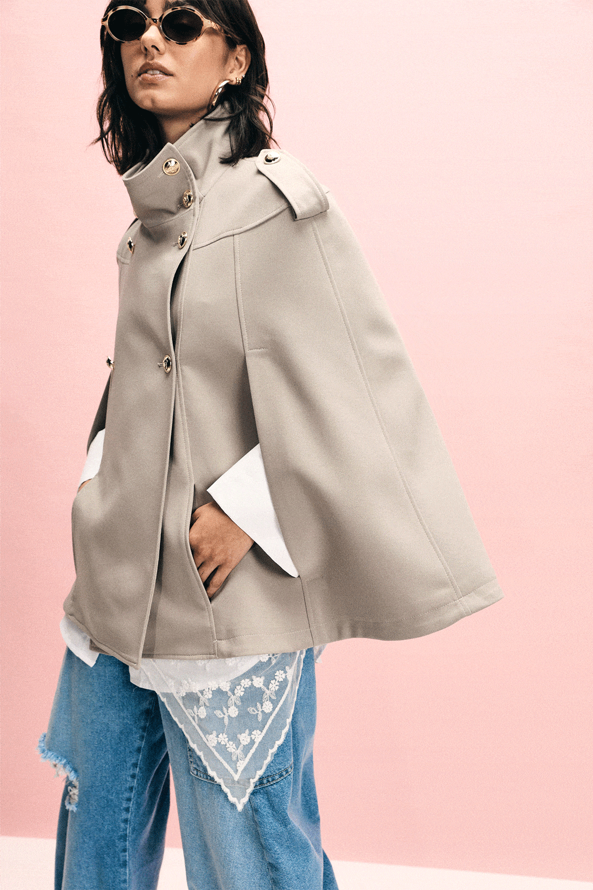 Fashion collage of a female model in sunglasses wearing a beige cape, oversized white shirt and distressed wide-leg jeans against a pink backdrop