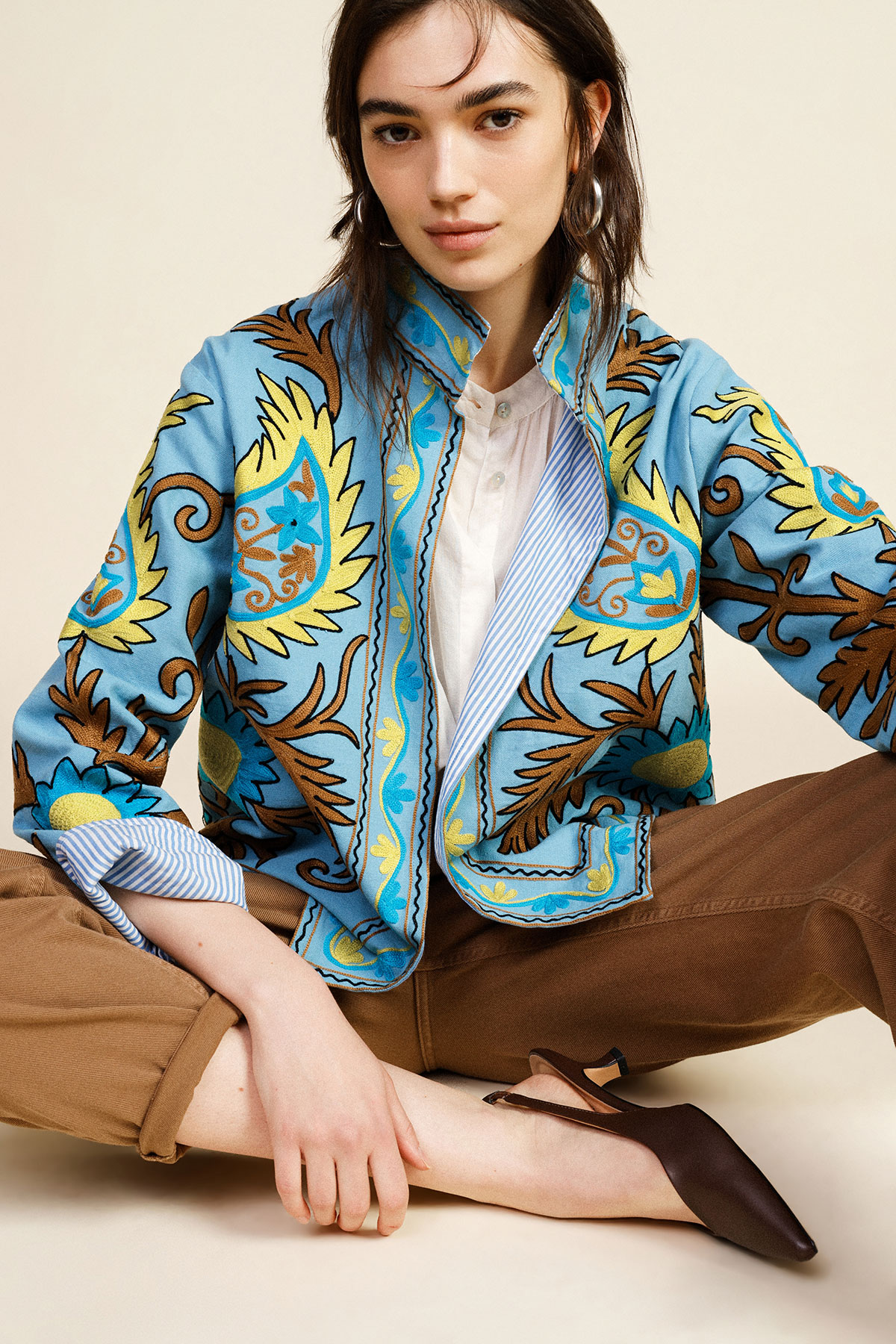 Young woman seated cross‑legged wearing a blue embroidered floral jacket, white blouse, brown corduroy trousers and brown flats