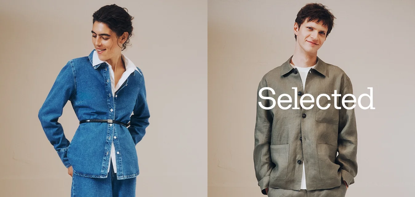 Due persone posano su uno sfondo semplice indossando abiti casual: una camicia di jeans con cintura e pantaloni, e una giacca verde con bottoni abbinata a pantaloni coordinati.