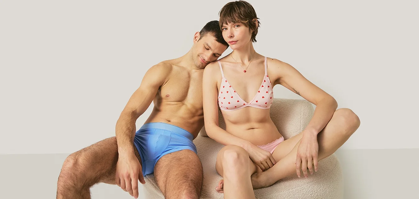 Uomo in costume da bagno blu che appoggia la testa sulla donna che indossa un bikini rosa a pois, entrambi seduti contro uno sfondo chiaro e uniforme.